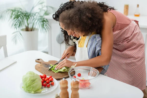 Joven madre afroamericana cortando repollo cerca de niños y tomates cherry en la cocina - foto de stock