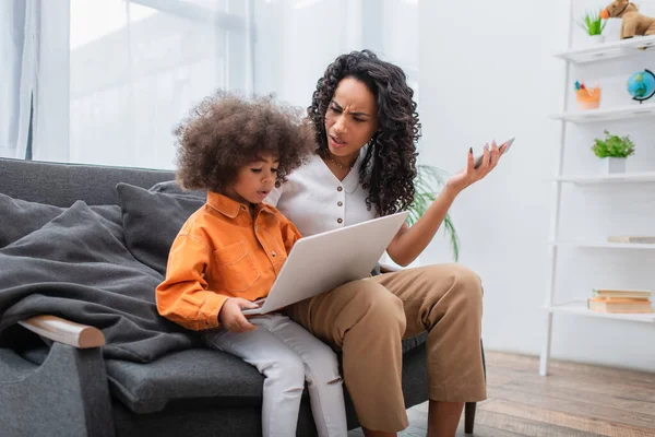 Angry african american woman holding smartphone near toddler kid using laptop on couch — Stock Photo