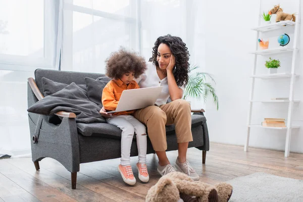 African american mother looking at daughter with laptop on couch in living room — Stock Photo