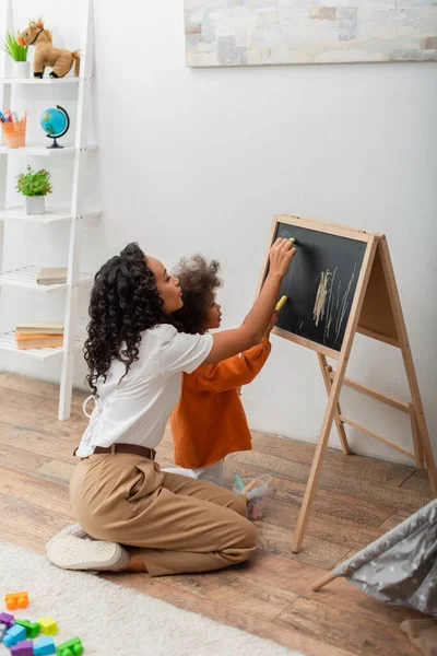 Vista lateral de la mujer afroamericana y el niño dibujo en pizarra en la sala de estar - foto de stock
