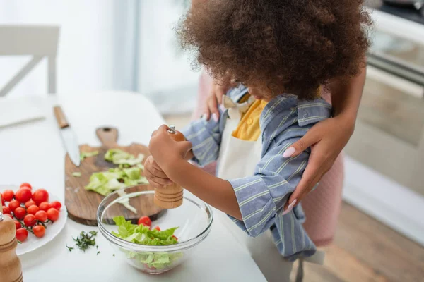 Niño afroamericano chica condimento ensalada cerca de mamá en cocina - foto de stock