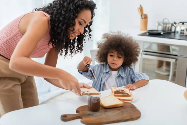 Sorridente donna afroamericana in possesso di pane vicino figlia e pasta di cioccolato in cucina — Foto stock