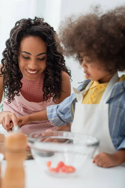 Positive african american mother cooking near blurred daughter in apron in kitchen — Stock Photo