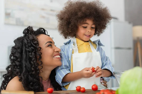 Mujer afroamericana feliz mirando al niño sosteniendo tomate cherry en la cocina - foto de stock