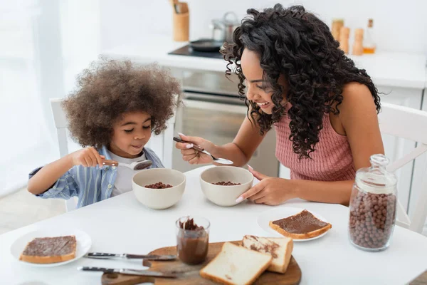 Madre e hija afroamericanas positivas sentadas cerca de cereales y pasta de chocolate durante el desayuno en casa - foto de stock