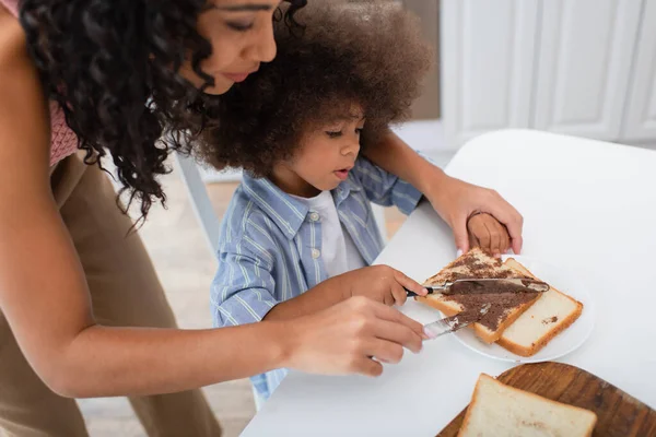 Mujer afroamericana y niño extendiendo pasta de chocolate en el pan en la cocina - foto de stock