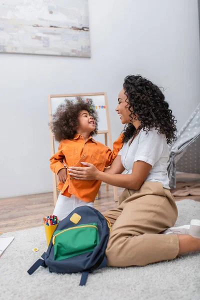 Lächelndes afrikanisch-amerikanisches Kind sieht Mama in der Nähe von Rucksack und Buntstiften zu Hause an — Stockfoto