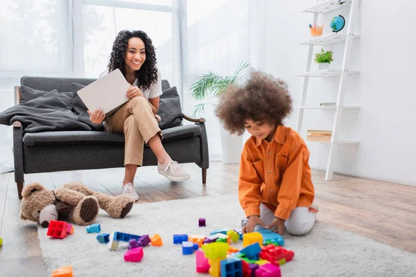 Sourire femme afro-américaine tenant un ordinateur portable près de la fille avec des blocs de construction à la maison — Photo de stock