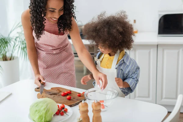 Sonriente mujer afroamericana sosteniendo cuchillo cerca de verduras frescas e hija en la cocina - foto de stock