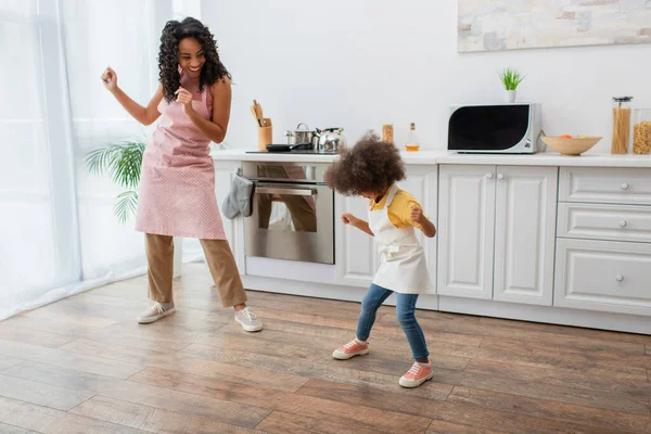Mujer afroamericana feliz y niño en delantales bailando en la cocina - foto de stock