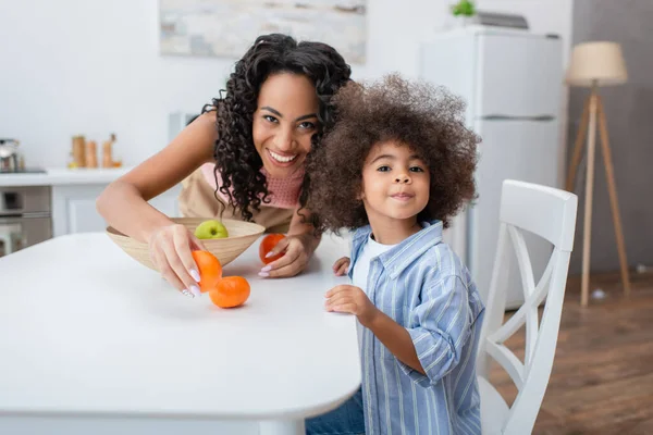 Positive african american woman holding fresh fruits near daughter in kitchen — Stock Photo