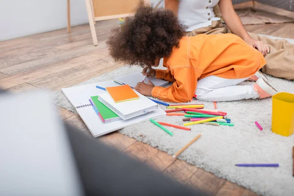 Afroamericano ragazza disegno su notebook vicino a matite di colore e madre a casa — Foto stock