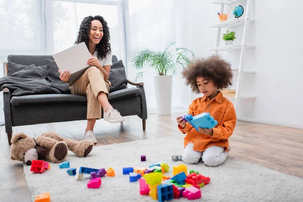 Autónomo afroamericano positivo mirando a un niño pequeño jugando bloques de construcción en casa - foto de stock