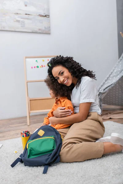 Heureuse femme afro-américaine étreignant sa fille près de crayons de couleur et sac à dos à la maison — Photo de stock