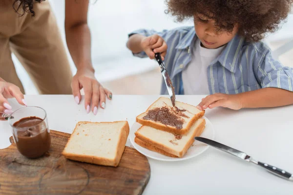 Африканская американка, стоящая рядом с дочерью, разбрасывает шоколадную пасту на хлеб — стоковое фото