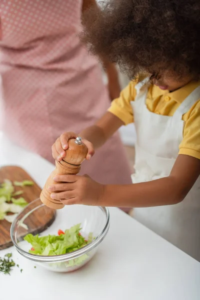 Africano menina americana tempero salada perto de mãe turva na cozinha — Fotografia de Stock
