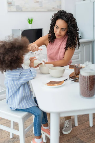 Jeune mère afro-américaine versant du lait près de sa fille et petit déjeuner dans la cuisine — Photo de stock