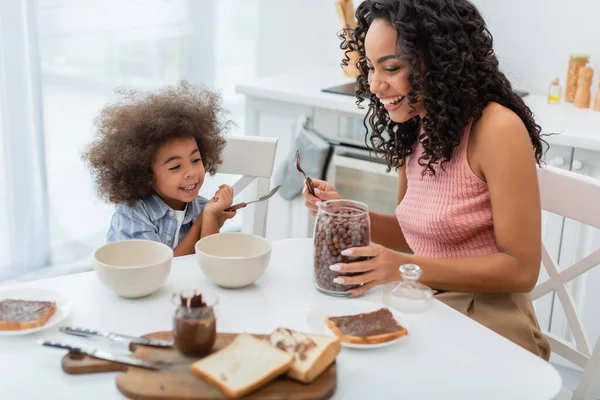 Glückliche afrikanisch-amerikanische Mutter und Kind sitzen neben Getreide und Brot mit Schokoladenpaste in der Küche — Stockfoto