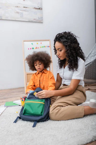 African american woman holding backpack near daughter with notebook and stationery at home — Stock Photo