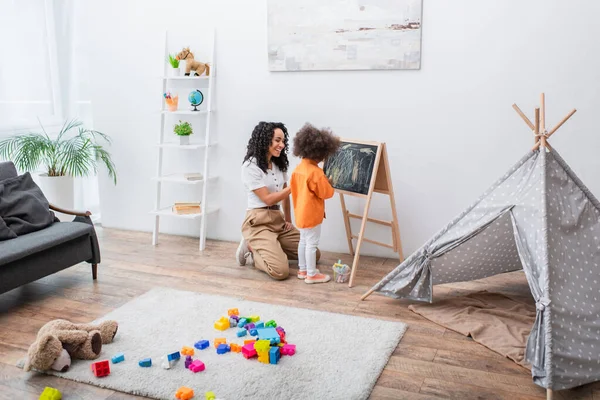 African american parent looking at kid with chalk near chalkboard and tent at home — Stock Photo