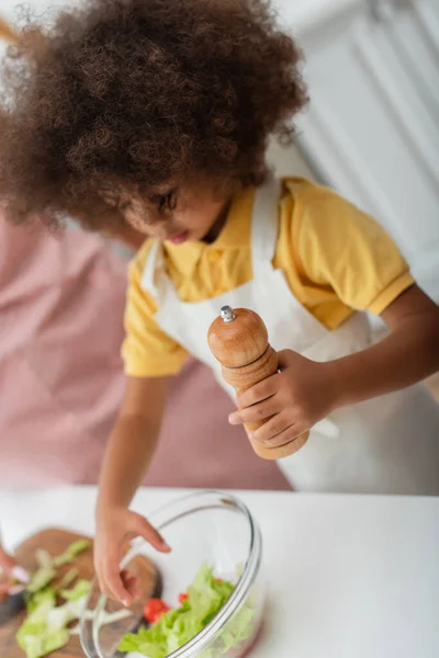 African american kid in apron holding salt mill near salad at home — Stock Photo