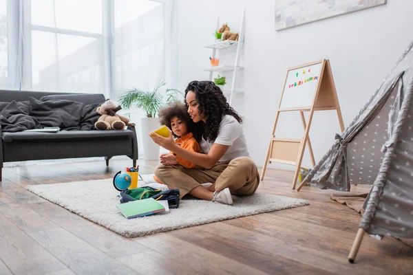 Mamma afroamericana che tiene il taccuino vicino a bambino, cancelleria e globo su tappeto a casa — Foto stock