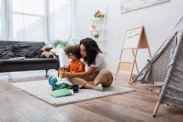 Joven afroamericana madre e hija sosteniendo lápices de color cerca de papelería y tienda de campaña en casa - foto de stock