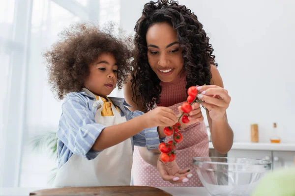 Positive african american mother and kid holding cherry tomatoes near cutting board in kitchen — Stock Photo