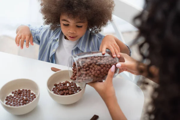 Afroamerikanerin gießt Müsli in Schüsseln mit Milch neben Kind in Küche — Stockfoto