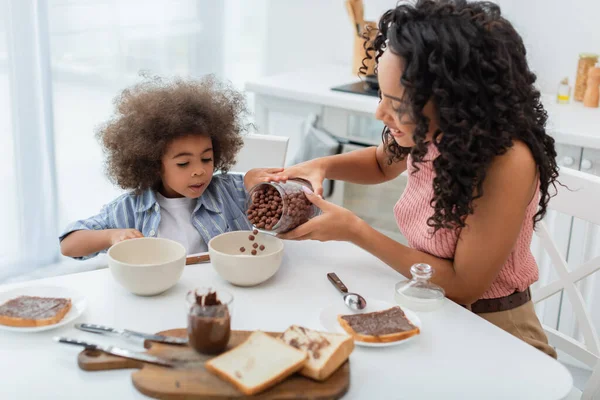 African american woman pouring cereal in bowl near bread and child in kitchen — Stock Photo