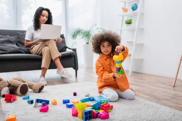 Africano criança americana jogando blocos de construção perto mãe turva com laptop em casa — Fotografia de Stock