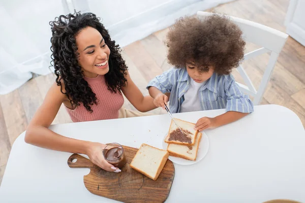 High angle view of smiling african american woman holding chocolate paste near kid with bread at home — Stock Photo