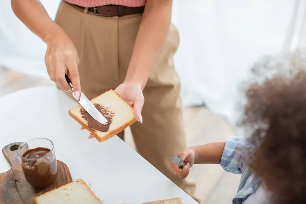 Vista ritagliata della donna afro-americana che diffonde pasta di cioccolato sul pane vicino al bambino sfocato — Foto stock