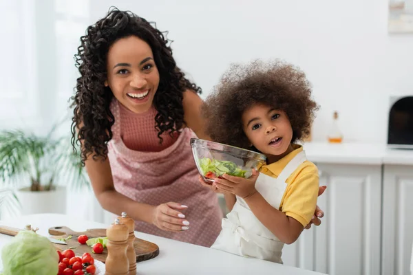 Sonriente mujer ameriana africana mirando a la cámara cerca de la hija con un tazón de ensalada en casa - foto de stock