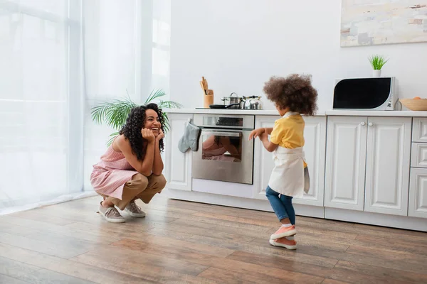 Lächelnde afrikanisch-amerikanische Mutter schaut Tochter in Schürze in Küche an — Stockfoto