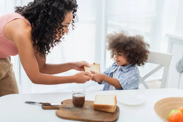 Sorrindo mulher afro-americana e criança segurando pão perto de pasta de chocolate na cozinha — Fotografia de Stock