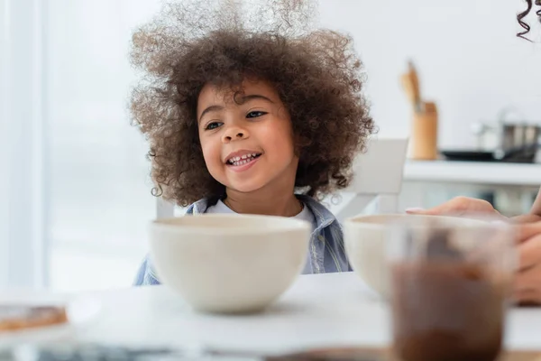 Lächelndes afrikanisches amerikanisches Kind sitzt neben Schüsseln und Mama in der Küche — Stockfoto