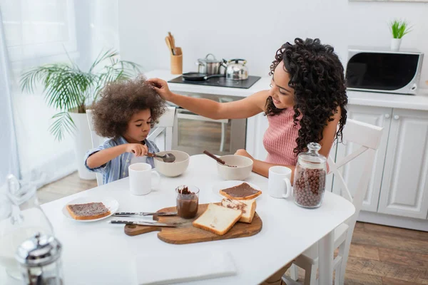 Sorridente donna afroamericana toccare bambino mangiare cereali vicino a colazione a casa — Foto stock