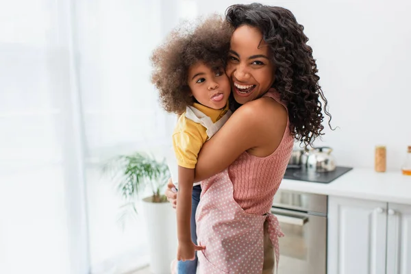 Heureuse mère afro-américaine dans un tablier étreignant l'enfant en train de sortir la langue dans la cuisine — Photo de stock