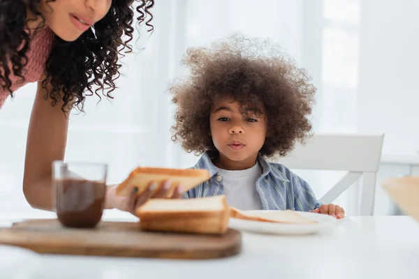 Niño afroamericano sentado cerca de mamá con pan y pasta de chocolate borrosa en la cocina - foto de stock