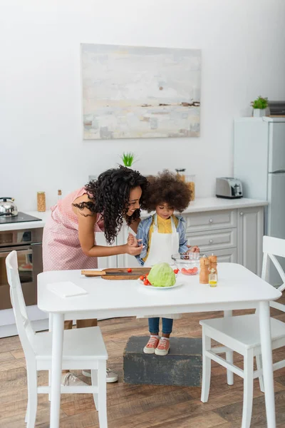 Sorridente donna afro-americana in piedi vicino a figlia e verdure fresche a casa — Foto stock