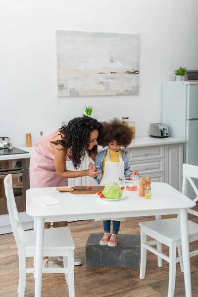 Sorrindo mulher afro-americana de pé perto da filha e legumes frescos em casa — Stock Photo