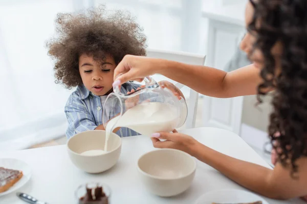 Blurred african american mother pouring milk in bowls near daughter in kitchen — Stock Photo