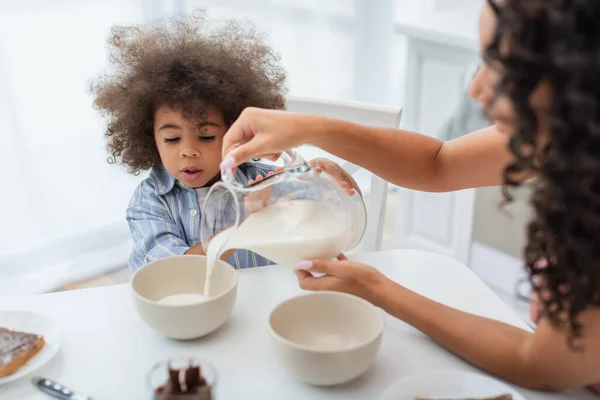 Borrosa africana americana madre verter leche en cuencos cerca hija en cocina - foto de stock