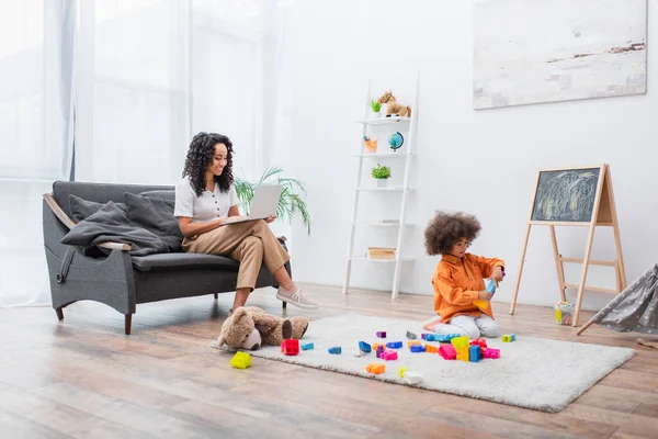 Lächelnde Frau mit Laptop auf der Couch, während Tochter zu Hause Bauklötze spielt — Stockfoto