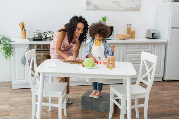 Lächelnde Afroamerikanerin steht neben Tochter mit Kirschtomaten und Schneidebrett in Küche — Stockfoto