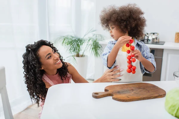 Africano americano chica holding cherry tomates cerca sonriendo mamá en casa - foto de stock