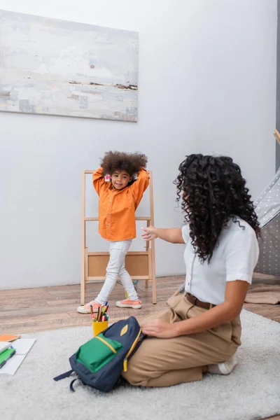 Sonriente niño afroamericano de pie cerca de la placa magnética y mamá con mochila en casa - foto de stock