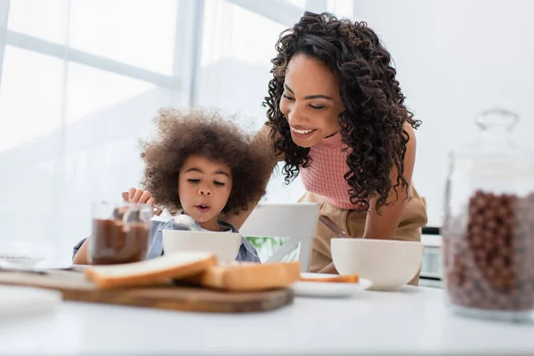 Smiling african american parent standing near daughter eating breakfast near cereal and chocolate paste — Stock Photo