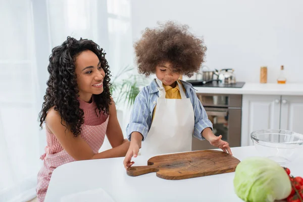 Positiva madre afroamericana en delantal de pie cerca de su hija, tabla de cortar y verduras frescas en casa - foto de stock